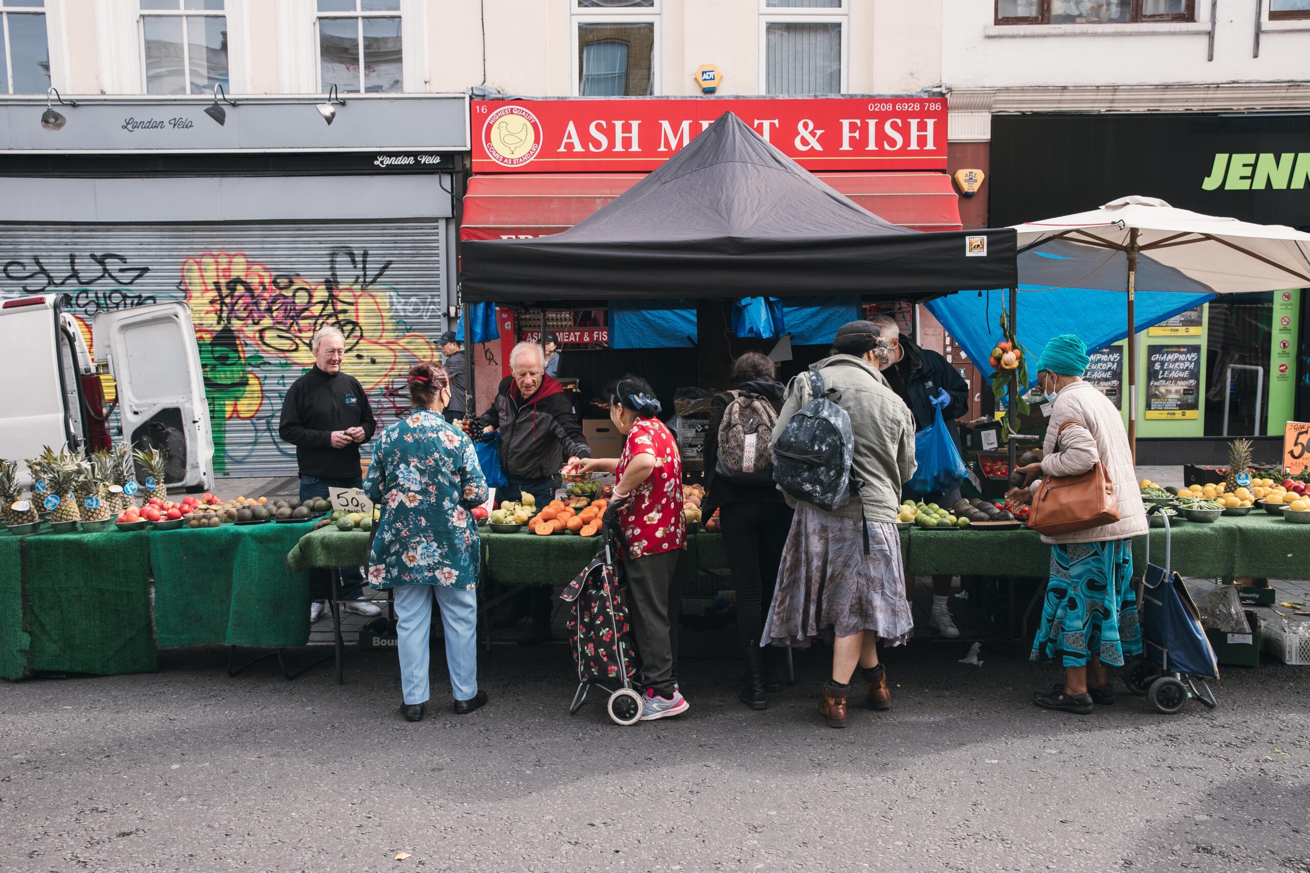 Image of a local market in London, showing a diverse group of people from behind shopping for fruit and vegetables.