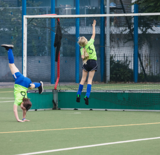 Two school girls dressed in sport kit on an outside football pitch; one is leaping to tap the top of a goal frame and the other, doing a cartwheel.