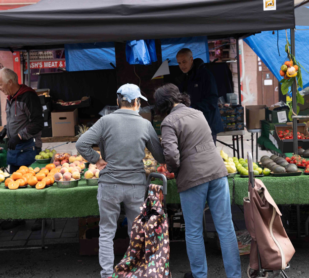 Two people stand at an outdoor market stall selecting fruits and vegetables from a display, with a vendor behind the table and other shoppers nearby.