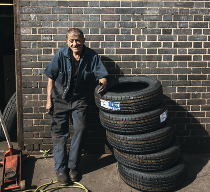 Work garage man An older man wearing dirtied overalls from a garage smiles at the camera, while leaning on a stack of black tyres.