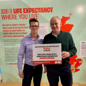 Leader of the Liberal Democrats Ed Davey MP holding a pledge board, committing to make health equal alongside Health Equals' Chief Campaigns Officer